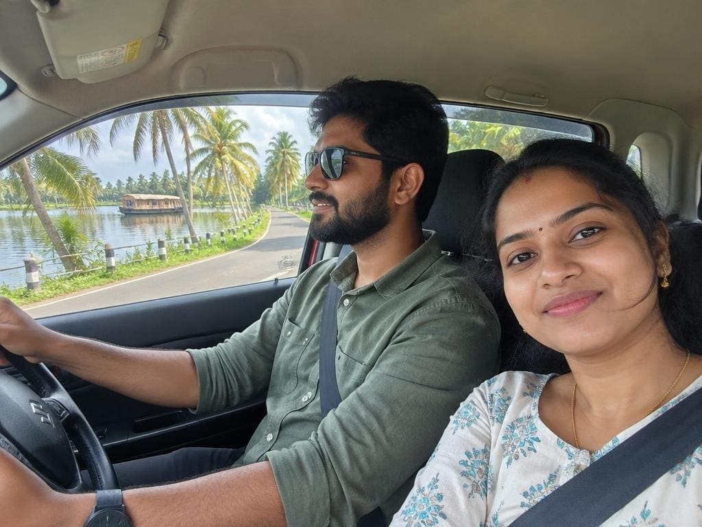 Couple in a car driving along a palm-lined backwater road near Alleppey with a houseboat on the water